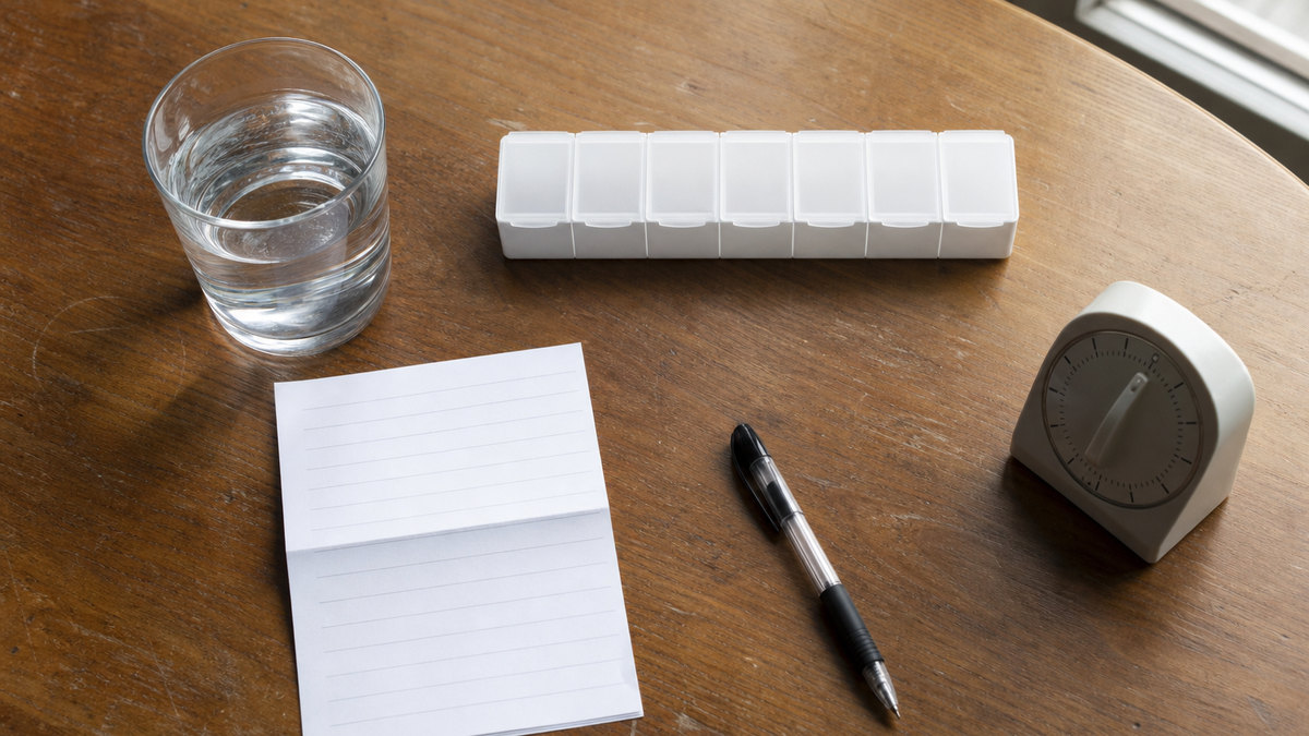 Naturaleza muerta editorial con un organizador semanal de pastillas, un vaso de agua y materiales sin marca para revisar medicamentos